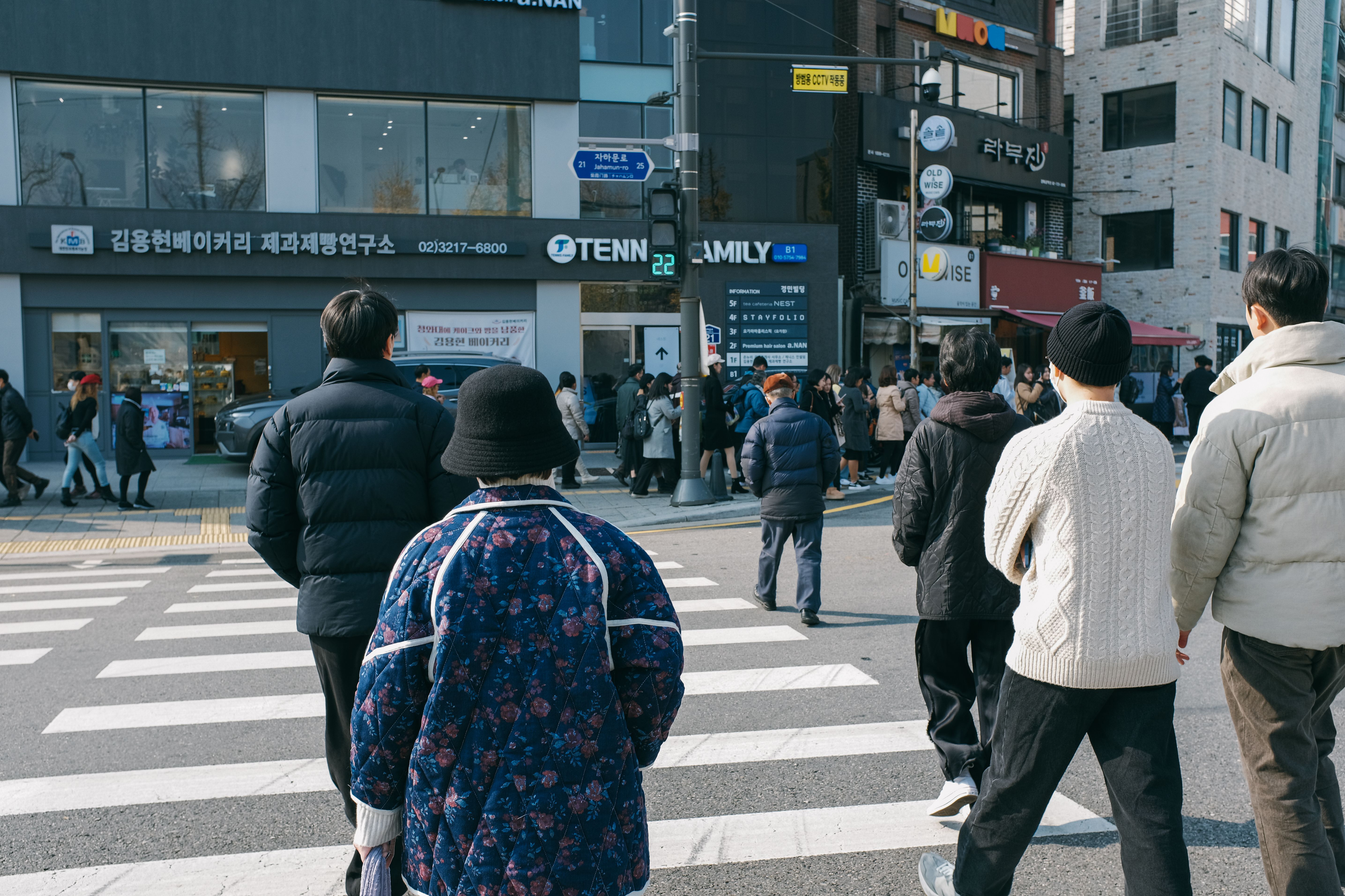 활기찬 도시 거리 풍경. 사람들이 걷거나 모여 앉아 분위기를 즐기고 있으며, 은은하게 음악이 들리는 듯한 느낌을 준다. 다채로운 색감의 도시 건축물과 사람들이 어우러져 역동적인 분위기를 연출한다.